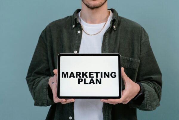 A man in casual attire displays a tablet with 'Marketing Plan' text against a blue background.